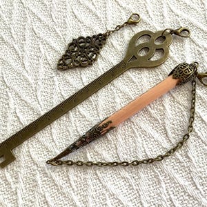 Pode incluir: Tr&ecirc;s marcadores de metal em estilo antigo e um berloque de l&aacute;pis com corrente. Os marcadores incluem uma chave, um formato de losango e um design de r&eacute;gua. O berloque de l&aacute;pis &eacute; rosa e tem uma ponta de metal decorativa.