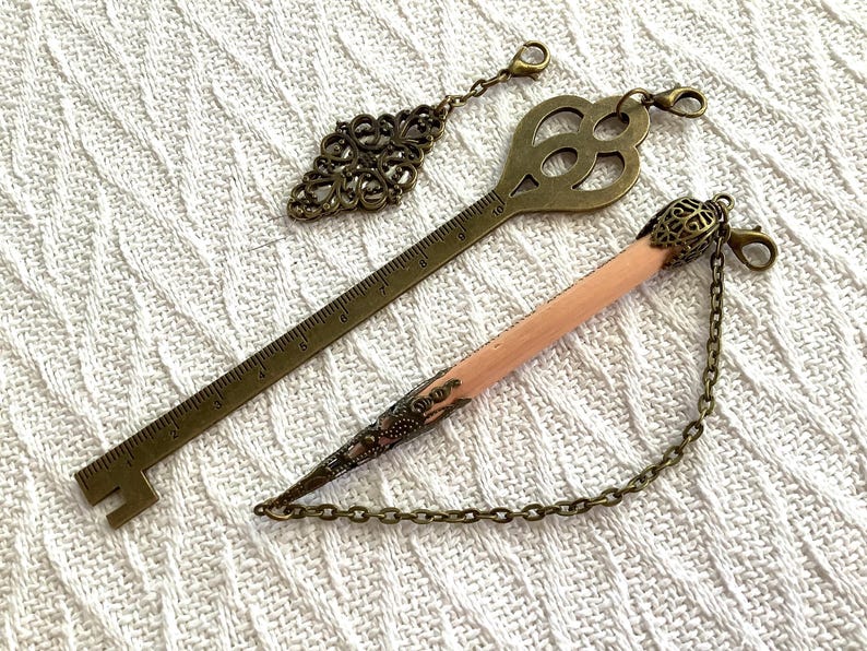 Pode incluir: Tr&ecirc;s marcadores de metal em estilo antigo e um berloque de l&aacute;pis com corrente. Os marcadores incluem uma chave, um formato de losango e um design de r&eacute;gua. O berloque de l&aacute;pis &eacute; rosa e tem uma ponta de metal decorativa.