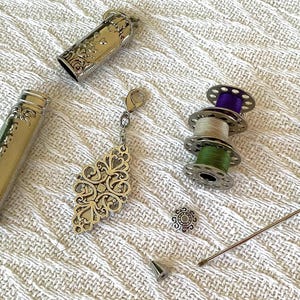 May include: Silver sewing kit with ornate floral designs. Includes a needle case, thimble, thread spools with purple, beige, and green thread, a decorative pendant, and a needle threader. The items are arranged on a white textured surface.