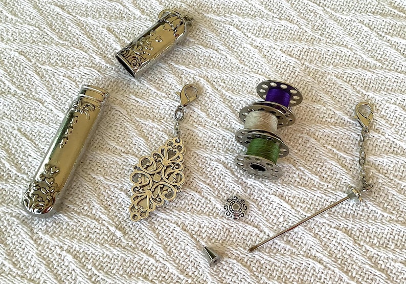 May include: Silver sewing kit with ornate floral designs. Includes a needle case, thimble, thread spools with purple, beige, and green thread, a decorative pendant, and a needle threader. The items are arranged on a white textured surface.