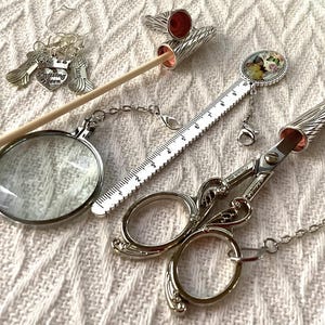 May include: A collection of silver knitting accessories, including scissors, a magnifying glass, a ruler, and stitch markers. The scissors have a decorative wing design. The stitch markers have charms that say "Knitting Queen".