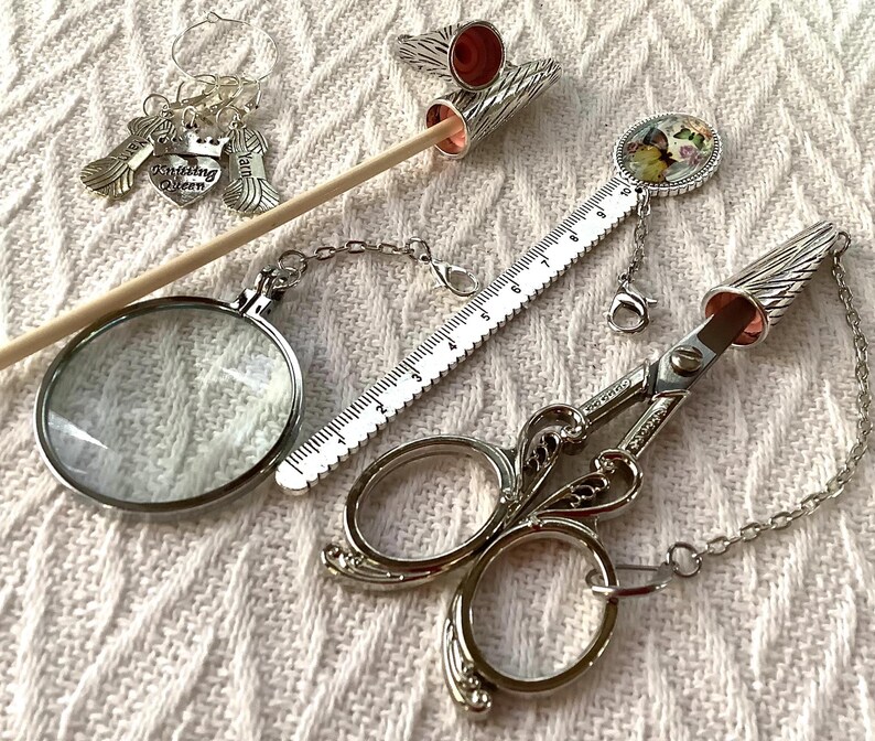 May include: A collection of silver knitting accessories, including scissors, a magnifying glass, a ruler, and stitch markers. The scissors have a decorative wing design. The stitch markers have charms that say "Knitting Queen".