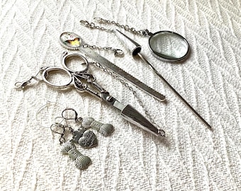 May include: A collection of silver-toned crochet tools and accessories. Includes scissors, a magnifying glass, a ruler, a yarn needle, and charms that say "Yarn" and "Crochet Queen". The items are arranged on a white, textured surface.