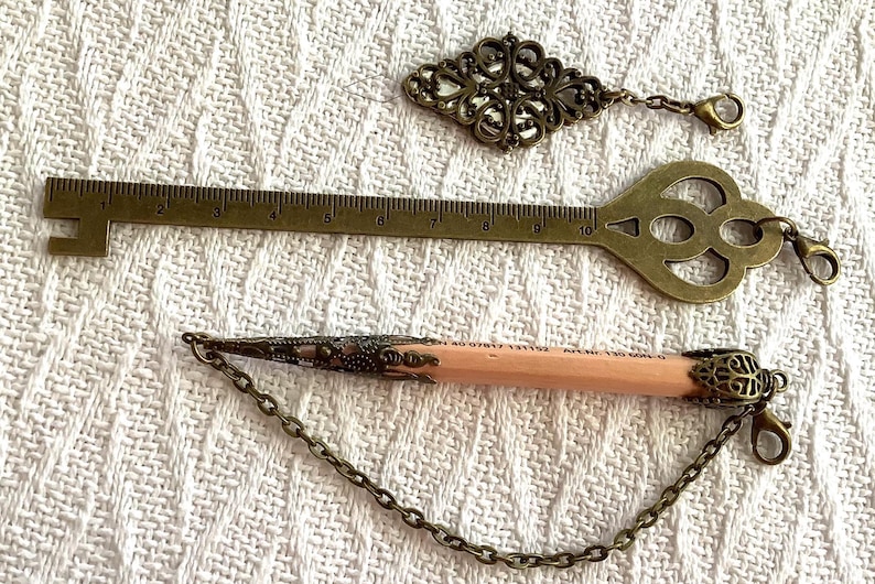 Pode incluir: Uma variedade de acess&oacute;rios antigos em tom de lat&atilde;o, incluindo uma r&eacute;gua em forma de chave, um pingente decorativo e um l&aacute;pis com extremidades met&aacute;licas decorativas. A r&eacute;gua em forma de chave mede aproximadamente 25 cm de comprimento. Todos os itens t&ecirc;m uma est&eacute;tica vintage.
