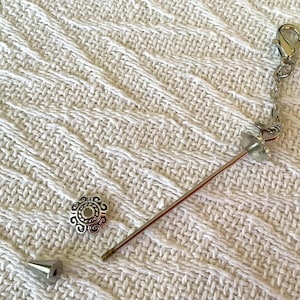 May include: A silver-toned bottle stopper with a decorative floral bead, a silver chain, and a metal pin. The stopper is accompanied by a decorative silver bead and a silver cone-shaped piece. The items are on a white textured surface.