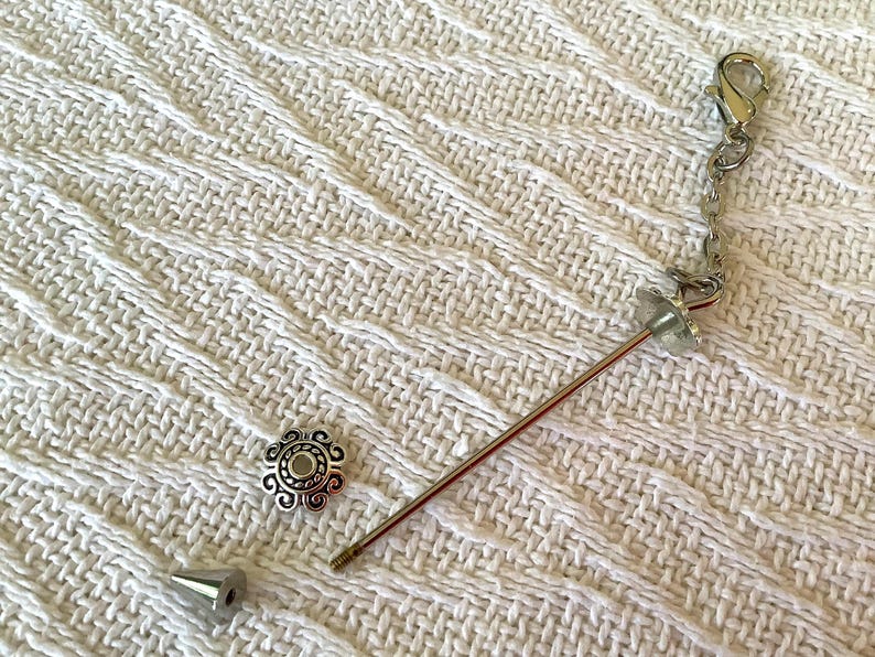 May include: A silver-toned bottle stopper with a decorative floral bead, a silver chain, and a metal pin. The stopper is accompanied by a decorative silver bead and a silver cone-shaped piece. The items are on a white textured surface.