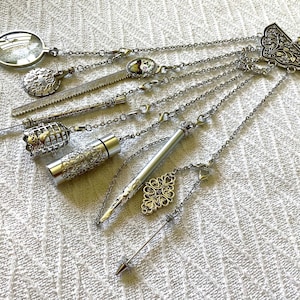 May include: A silver-toned multi-chain accessory featuring a decorative fan-shaped top piece. Attached chains hold various charms including a magnifying glass, a textured metal disc, a small birdcage, a cylindrical container, and a pointed stylus.