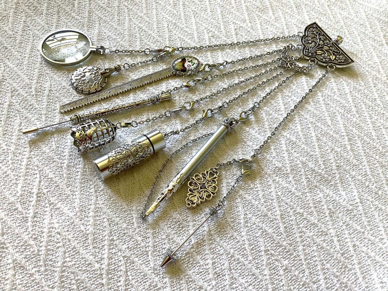 May include: A silver-toned multi-chain accessory featuring a decorative fan-shaped top piece. Attached chains hold various charms including a magnifying glass, a textured metal disc, a small birdcage, a cylindrical container, and a pointed stylus.