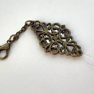 May include: An ornate, antique brass-coloured charm with a decorative filigree design. The charm is diamond-shaped with a small chain and clasp attached. A thin, clear thread extends from the charm.