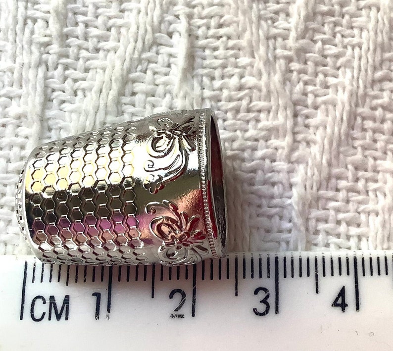 May include: Silver thimble with a hexagonal pattern and floral design. The thimble is approximately 1.5 inches in diameter.