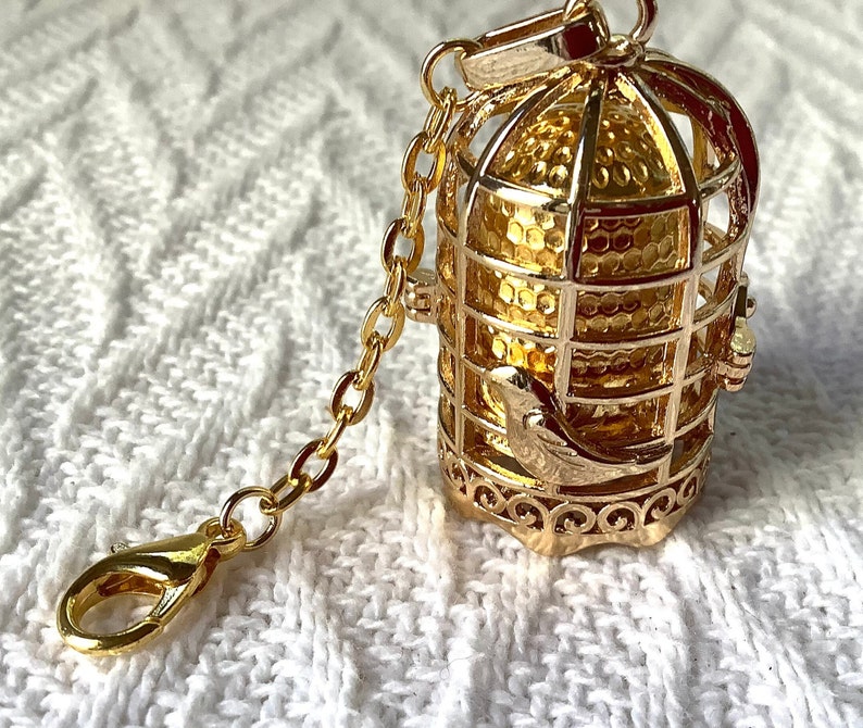 May include: A gold-tone birdcage charm with a small bird inside. The cage is attached to a gold-tone chain with a lobster clasp.
