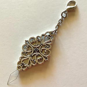 May include: A silver-colored needle threader with an ornate, filigree design. The threader has a decorative, diamond-shaped body with a small needle extending from the bottom. A chain and clasp are attached to the top.