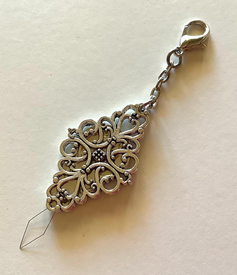 May include: A silver-colored needle threader with an ornate, filigree design. The threader has a decorative, diamond-shaped body with a small needle extending from the bottom. A chain and clasp are attached to the top.