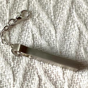 May include: A silver-colored keychain with a rectangular bar and a chain attached to a clasp. The keychain is made of metal and is resting on a white, textured surface.