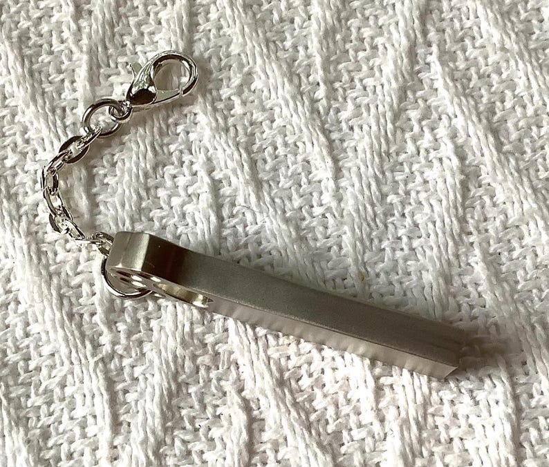 May include: A silver-colored keychain with a rectangular bar and a chain attached to a clasp. The keychain is made of metal and is resting on a white, textured surface.