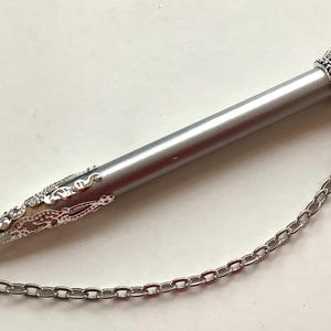 May include: A silver-toned cylindrical pen with a pointed, ornate filigree tip and a crown-shaped cap. A silver metal chain connects the cap to the tip, and a lobster claw clasp is attached to the top of the crown-shaped cap.