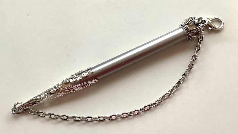 May include: A silver-toned cylindrical pen with a pointed, ornate filigree tip and a crown-shaped cap. A silver metal chain connects the cap to the tip, and a lobster claw clasp is attached to the top of the crown-shaped cap.