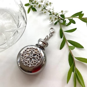 May include: A silver-coloured flask with a decorative filigree design on the front. The flask has a screw-on cap and a small chain with a clasp. The image also includes a cut-glass bowl, and sprigs of white flowers and green leaves.