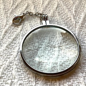 May include: A circular silver-toned metal locket frame with a clear glass front and a hinged closure. The locket features a small silver metal chain attached to a lobster claw clasp at the top, allowing it to be used as a charm or pendant.