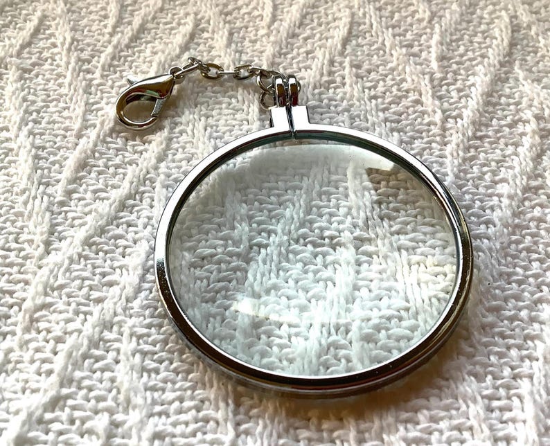 May include: A circular silver-toned metal locket frame with a clear glass front and a hinged closure. The locket features a small silver metal chain attached to a lobster claw clasp at the top, allowing it to be used as a charm or pendant.
