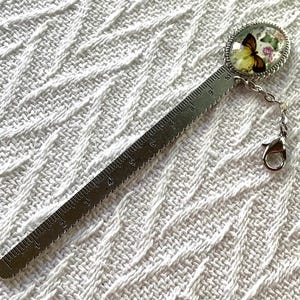 May include: A silver metal bookmark with a butterfly design. The bookmark features a ruler with inch measurements and a decorative oval charm with a butterfly and floral design. A small chain and clasp are attached to the charm.