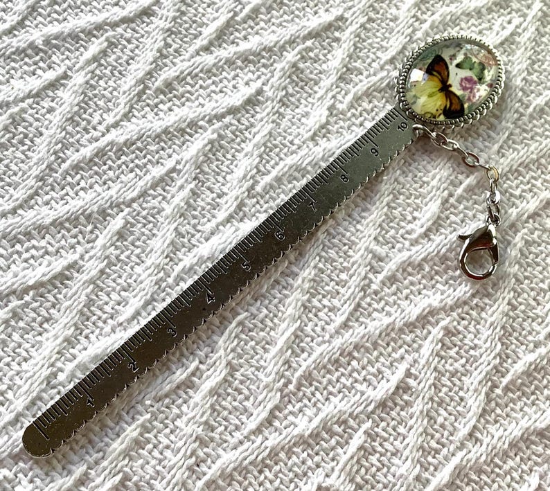 May include: A silver metal bookmark with a butterfly design. The bookmark features a ruler with inch measurements and a decorative oval charm with a butterfly and floral design. A small chain and clasp are attached to the charm.
