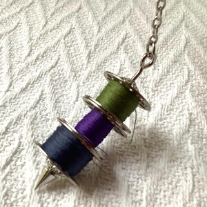 May include: A unique pendant featuring three spools of thread in green, purple, and navy blue. The spools are held together with silver-colored metal rings and a chain. The pendant has a silver-colored, pointed tip.
