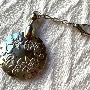 May include: A silver-toned circular charm featuring an embossed floral pattern with swirling details. The charm is attached to a short silver chain that ends in a lobster claw clasp, allowing it to be easily clipped onto various items or accessories.