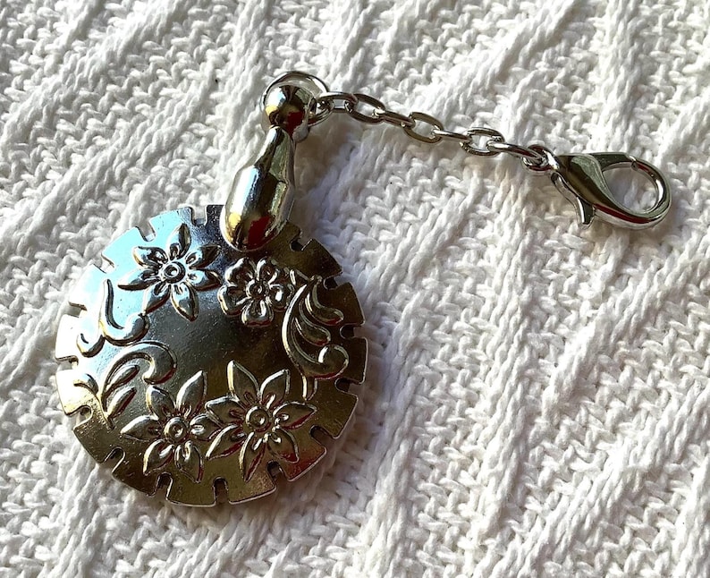 May include: A silver-toned circular charm featuring an embossed floral pattern with swirling details. The charm is attached to a short silver chain that ends in a lobster claw clasp, allowing it to be easily clipped onto various items or accessories.
