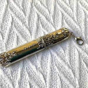 May include: A silver-toned needle case with ornate floral and scrollwork detailing. The cylindrical case has a hinged lid and a small clasp for attachment. The design is vintage-inspired, with a polished finish.