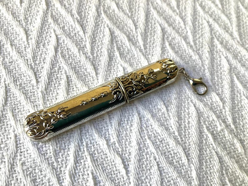 May include: A silver-toned needle case with ornate floral and scrollwork detailing. The cylindrical case has a hinged lid and a small clasp for attachment. The design is vintage-inspired, with a polished finish.