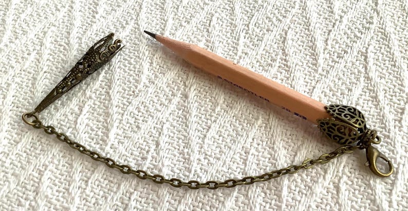 Pode incluir: Um l&aacute;pis de madeira com um porta-l&aacute;pis decorativo de cor bronze e uma corrente. O l&aacute;pis tem uma ponta de grafite afiada. O porta-l&aacute;pis tem um design de filigrana ornamentado. A corrente &eacute; presa a um fecho.