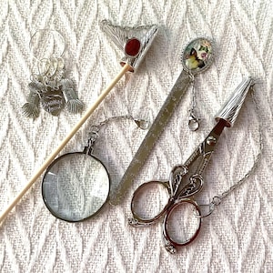 May include: A collection of silver-toned craft tools, including a pair of scissors, a magnifying glass, a ruler, and stitch markers. The scissors have a decorative handle and a chain. The ruler has a butterfly charm. The stitch markers have yarn ball charms.