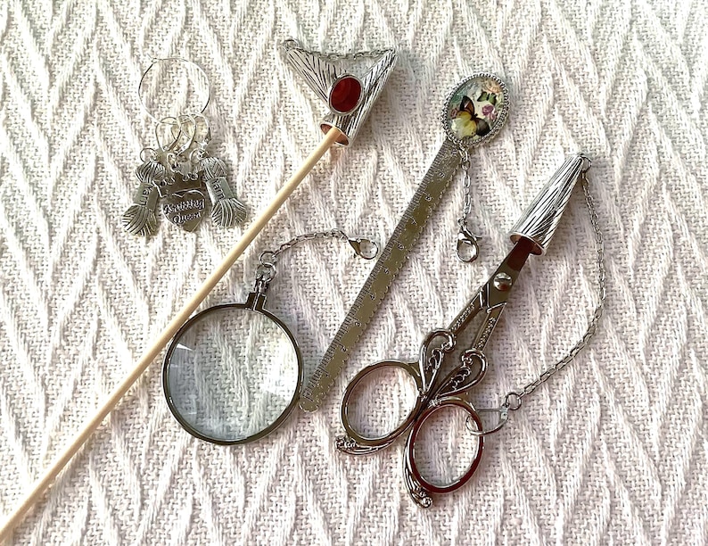 May include: A collection of silver-toned craft tools, including a pair of scissors, a magnifying glass, a ruler, and stitch markers. The scissors have a decorative handle and a chain. The ruler has a butterfly charm. The stitch markers have yarn ball charms.