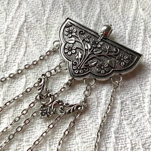 May include: A silver-toned metal brooch featuring a fan-shaped top piece with an embossed floral and leaf pattern. Multiple delicate silver chains hang from the base, with one central chain section incorporating an ornate filigree connector.
