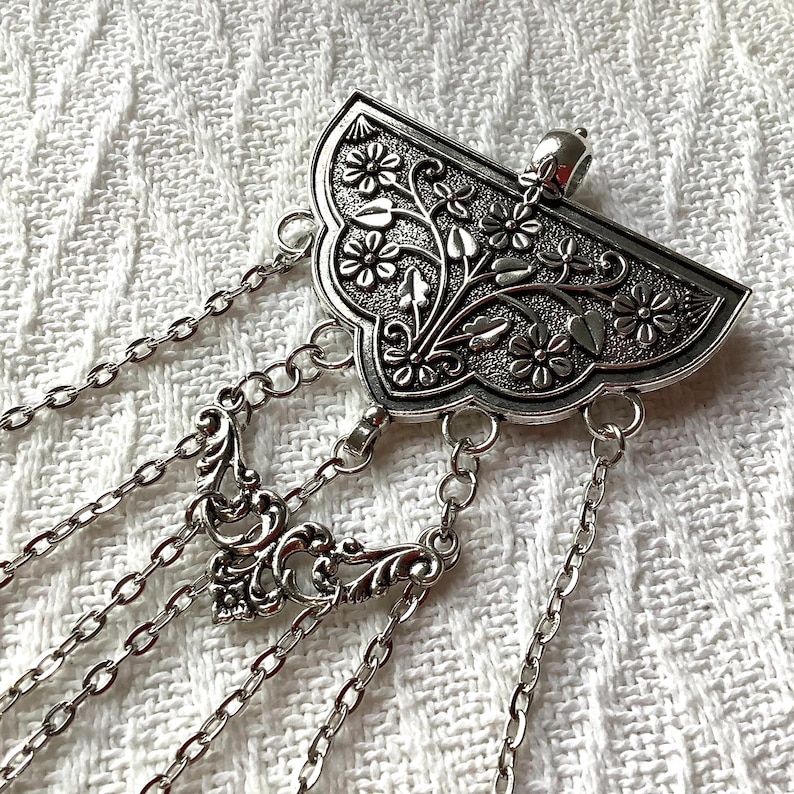 May include: A silver-toned metal brooch featuring a fan-shaped top piece with an embossed floral and leaf pattern. Multiple delicate silver chains hang from the base, with one central chain section incorporating an ornate filigree connector.