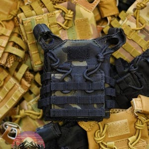 Puede incluir: Un chaleco t&aacute;ctico en miniatura negro y camuflaje con m&uacute;ltiples correas y un parche de velcro. El chaleco est&aacute; rodeado de otros chalecos en varios patrones y colores de camuflaje, creando una exhibici&oacute;n de tem&aacute;tica militar.