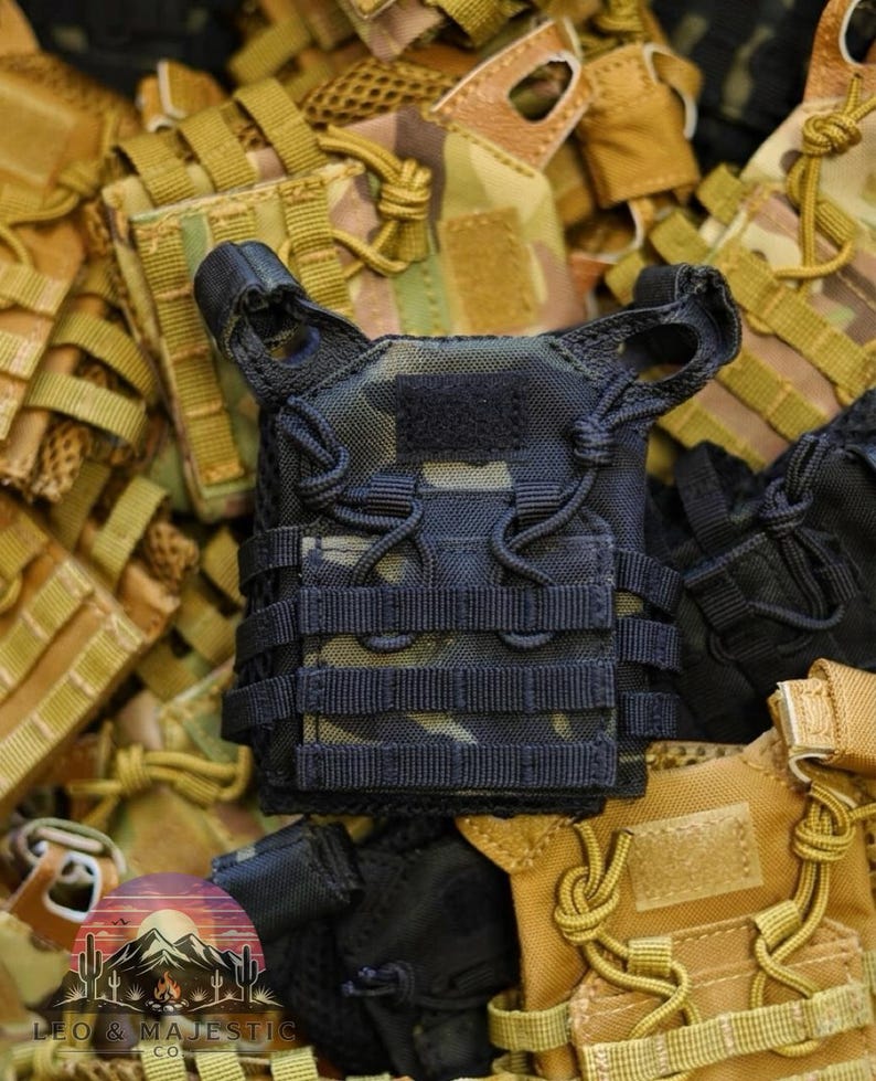 Puede incluir: Un chaleco t&aacute;ctico en miniatura negro y camuflaje con m&uacute;ltiples correas y un parche de velcro. El chaleco est&aacute; rodeado de otros chalecos en varios patrones y colores de camuflaje, creando una exhibici&oacute;n de tem&aacute;tica militar.