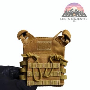 Puede incluir: Un chaleco t&aacute;ctico color canela con m&uacute;ltiples bolsillos y correas. El chaleco tiene un parche de velcro y est&aacute; sujeto por una mano enguantada. El logotipo "LEO & MAJESTIC CO." est&aacute; en la esquina superior derecha.