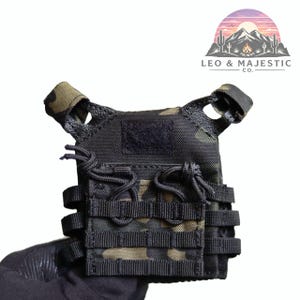 Puede incluir: Chaleco t&aacute;ctico negro y camuflaje con m&uacute;ltiples correas y un parche de velcro. El chaleco est&aacute; sujeto por una mano enguantada. El logotipo "LEO & MAJESTIC CO." es visible en la esquina superior derecha. Dise&ntilde;o robusto de inspiraci&oacute;n militar.