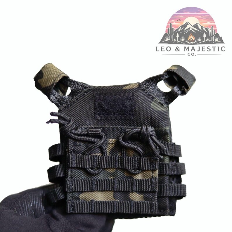 Puede incluir: Chaleco t&aacute;ctico negro y camuflaje con m&uacute;ltiples correas y un parche de velcro. El chaleco est&aacute; sujeto por una mano enguantada. El logotipo "LEO & MAJESTIC CO." es visible en la esquina superior derecha. Dise&ntilde;o robusto de inspiraci&oacute;n militar.