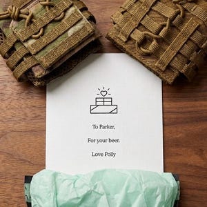 Puede incluir: Se muestran dos bolsas de equipo t&aacute;ctico en colores camuflaje y beige. Una tarjeta con una ilustraci&oacute;n de coraz&oacute;n y casa dice: &laquo;Para Parker, por tu cerveza. Love Polly&raquo;. Se ve una caja morada con papel de seda verde.
