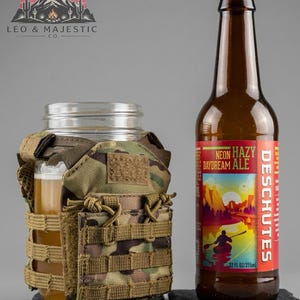 Puede incluir: Un tarro de cristal transparente con una funda de camuflaje que contiene un vaso de cerveza, junto a una botella de cerveza marr&oacute;n con una etiqueta roja y amarilla que dice "NEON HAZY DAYDREAM ALE". La etiqueta tambi&eacute;n dice "DESCHUTES" y "355ml". El logo "LEO & MAJESTIC CO." es visible.
