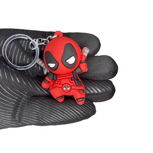 Peut inclure: Un porte-cl&eacute;s en caoutchouc repr&eacute;sentant le personnage de Deadpool en rouge et noir. Le porte-cl&eacute;s est en 3D et le personnage tient deux katanas.
