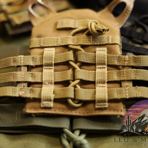 Puede incluir: Primer plano de un chaleco t&aacute;ctico color canela con m&uacute;ltiples correas y un dise&ntilde;o modular. El chaleco presenta una serie de correas tejidas y una bolsa central. La imagen tambi&eacute;n muestra un parche con un paisaje de monta&ntilde;a y desierto.