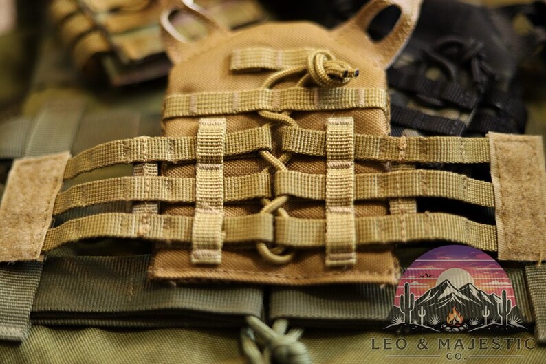 Puede incluir: Primer plano de un chaleco t&aacute;ctico color canela con m&uacute;ltiples correas y un dise&ntilde;o modular. El chaleco presenta una serie de correas tejidas y una bolsa central. La imagen tambi&eacute;n muestra un parche con un paisaje de monta&ntilde;a y desierto.