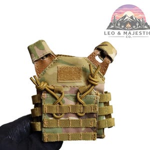 Puede incluir: Mini chaleco t&aacute;ctico de camuflaje en tonos verdes y beige. Cuenta con m&uacute;ltiples correas, un parche de velcro y cordones ajustables. El chaleco es sostenido por una mano enguantada. El logotipo "LEO & MAJESTIC CO." es visible.
