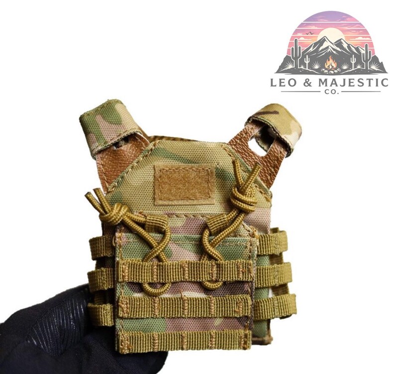 Puede incluir: Mini chaleco t&aacute;ctico de camuflaje en tonos verdes y beige. Cuenta con m&uacute;ltiples correas, un parche de velcro y cordones ajustables. El chaleco es sostenido por una mano enguantada. El logotipo "LEO & MAJESTIC CO." es visible.