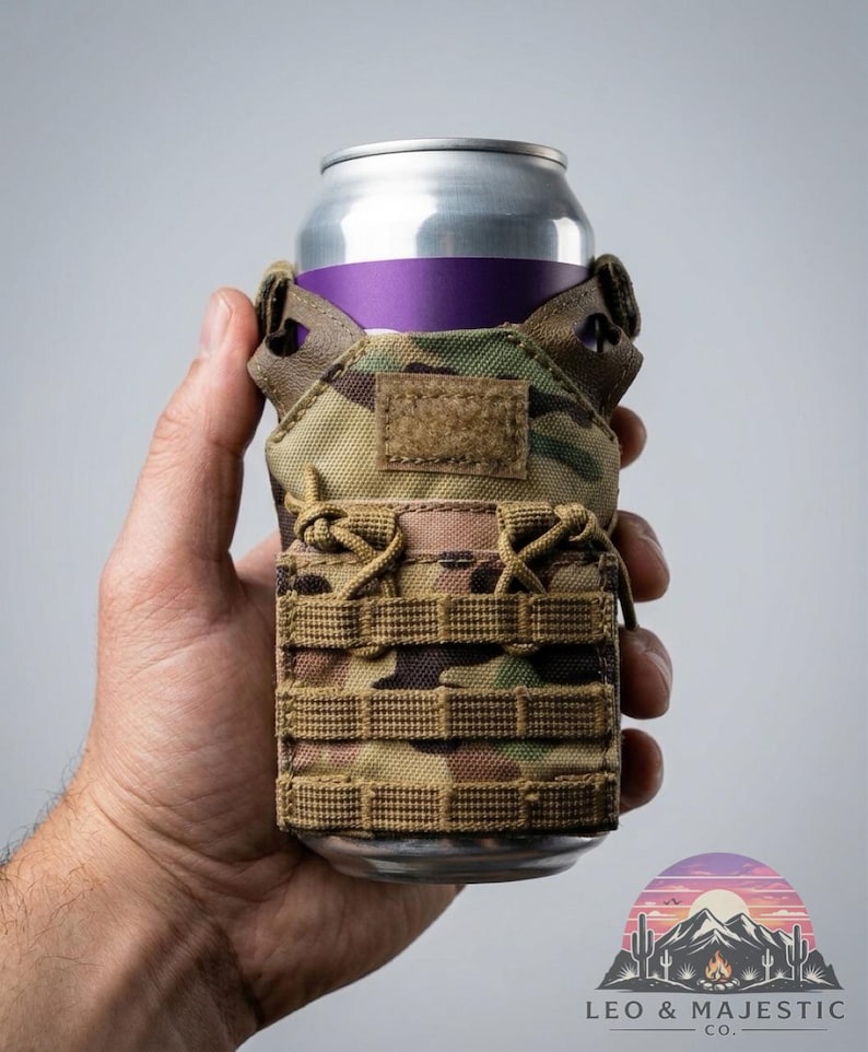 Puede incluir: Un enfriador de latas con estampado de camuflaje con un dise&ntilde;o t&aacute;ctico, sostenido en una mano. El enfriador presenta una rejilla de correas y un peque&ntilde;o parche. La lata interior tiene una banda morada. El logotipo "LEO & MAJESTIC CO." es visible.