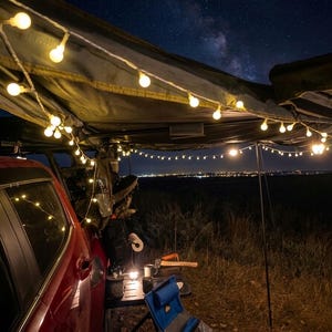 May include: A red vehicle with a camping setup under a dark canopy, illuminated by string lights. The night sky is visible with a view of the Milky Way. Camping gear includes a small table, chair, and a lantern.